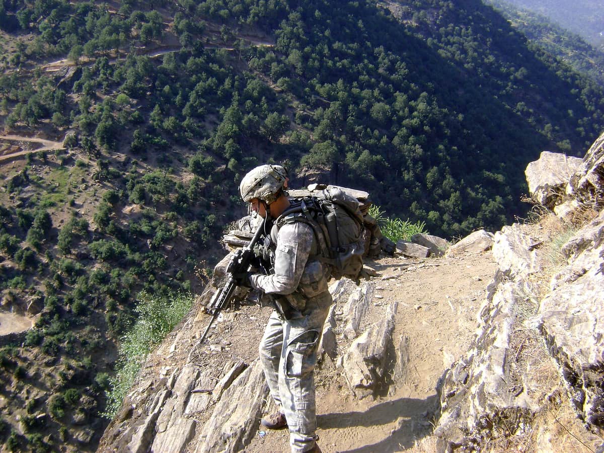 A soldier with a rifle and a heavy pack picking his way along a narrow rock spur high above a forested valley