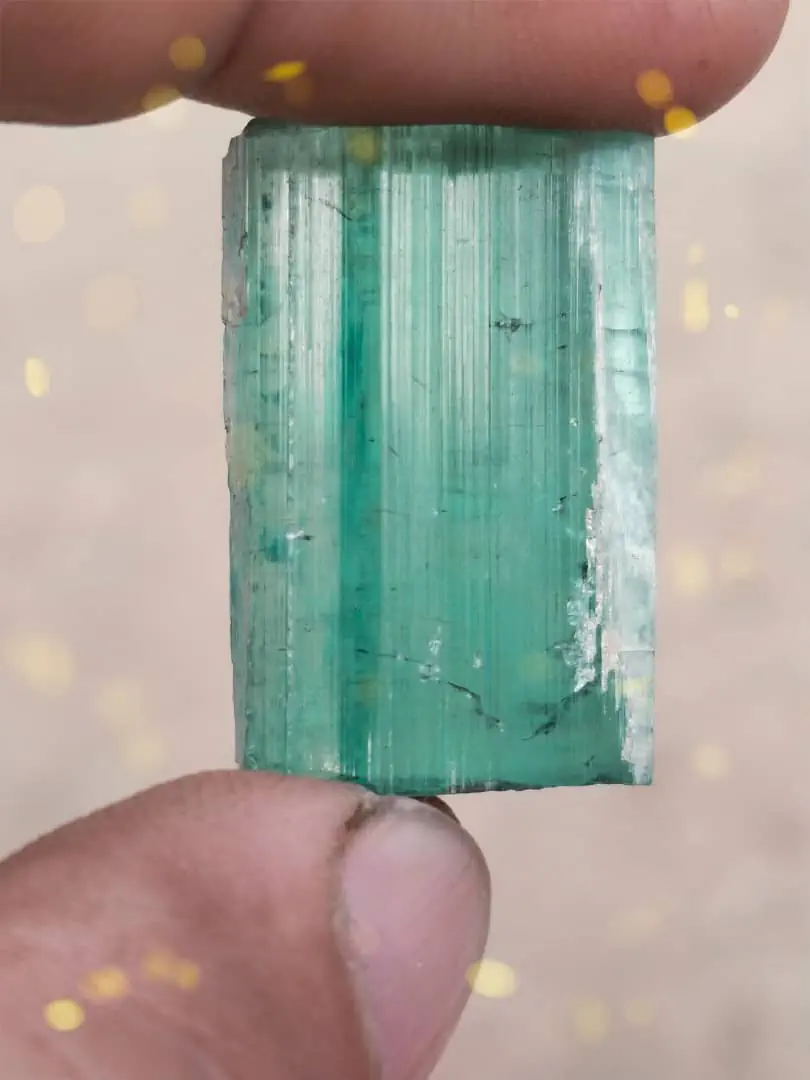 A blue-green tourmaline crystal from the Sewketra valley in Waygal, Nuristan, held up between finger and thumb, its faces striated along the length and a crust of white feldspar on one edge