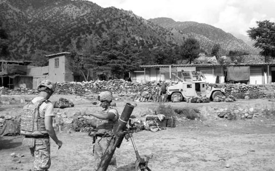 A black and white photograph of two soldiers at a mortar in an open field, a Humvee behind a low stone wall and the flat-roofed shops of the Wanat bazaar beyond, under a forested ridge