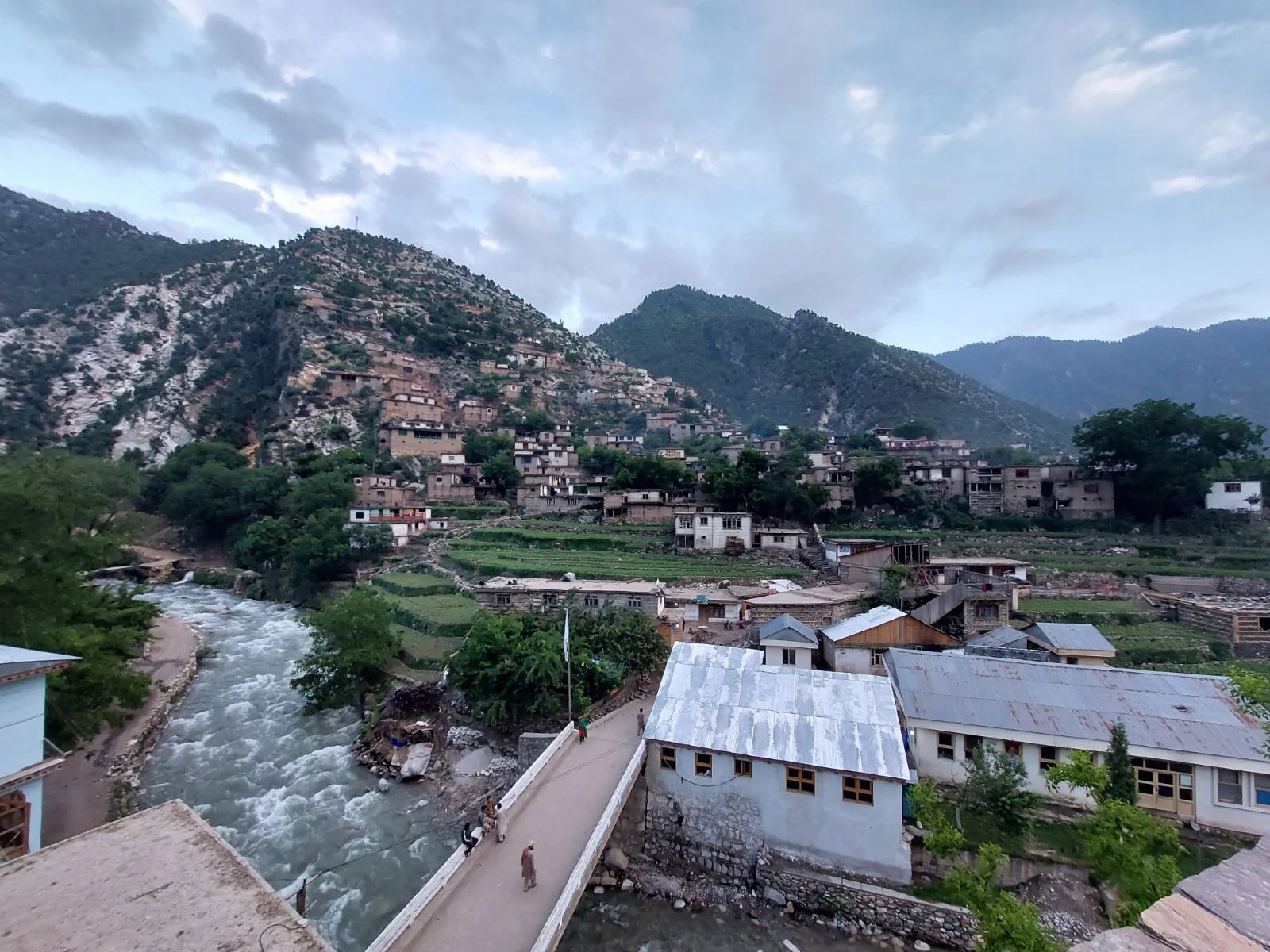 A bridge over the Waygal river at the district centre, people crossing, tin-roofed buildings beside it and the old village tiered up the hillside