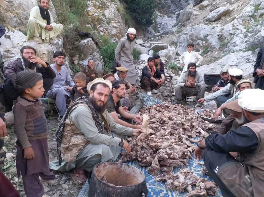 Men and boys of Waygal gathered on a mat on rocky ground around a large pile of goat meat being divided for a feast, a blackened cooking pot in the foreground