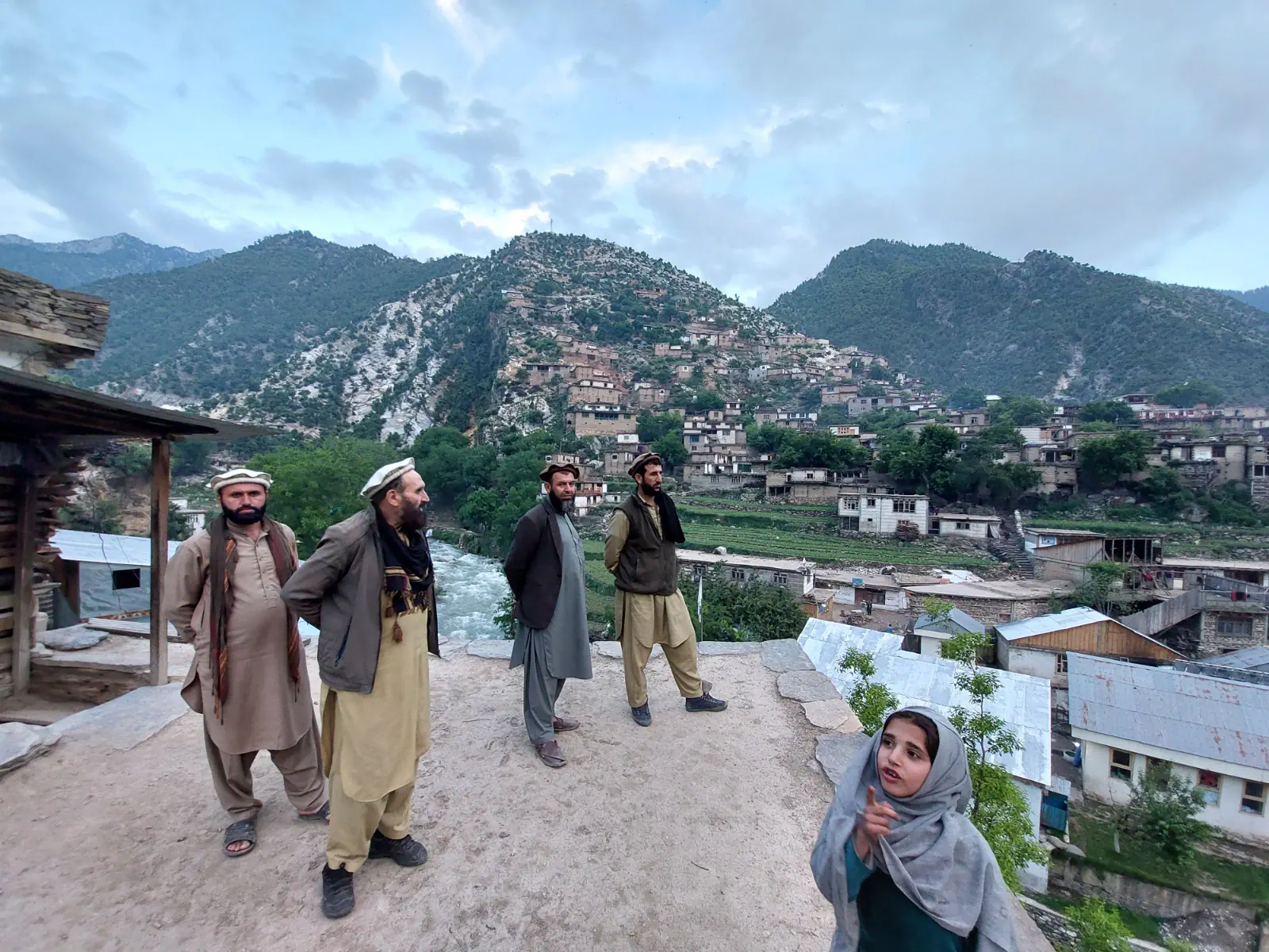 Four men in pakols and waistcoats on a rooftop above the centre of Waygal, a girl in a headscarf pointing in the foreground, the old village climbing the hill behind