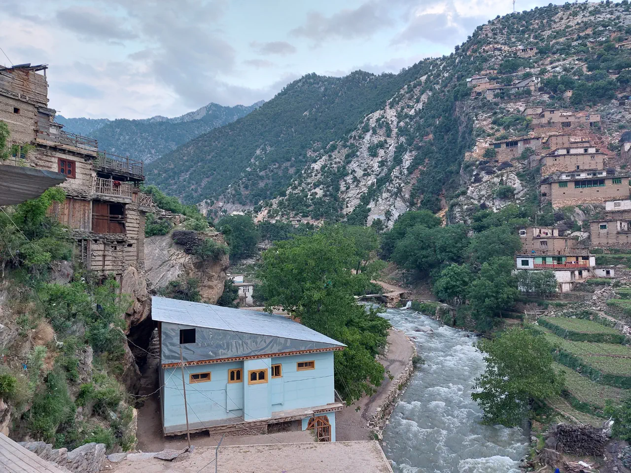 An old stone house laced with timber, with wooden balconies, beside a pale blue building on the Waygal river, tiered houses on the slope opposite