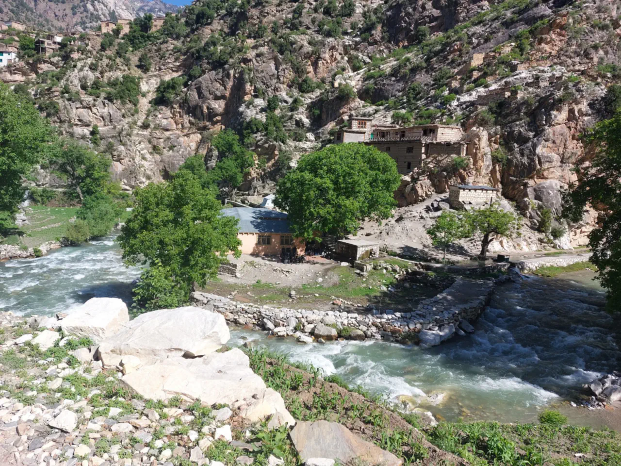 The Waygal river running fast past a stone weir and a blue-roofed building, old stone and timber houses on the cliffs above