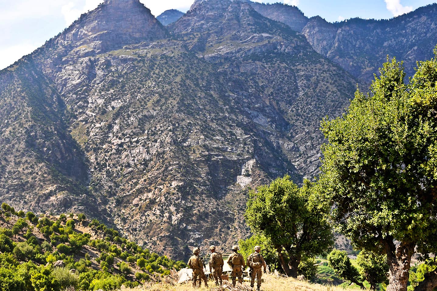 Four soldiers standing among holly oaks on a terraced slope, looking down into the Waygal valley at Wanat beneath a wall of bare rock mountains