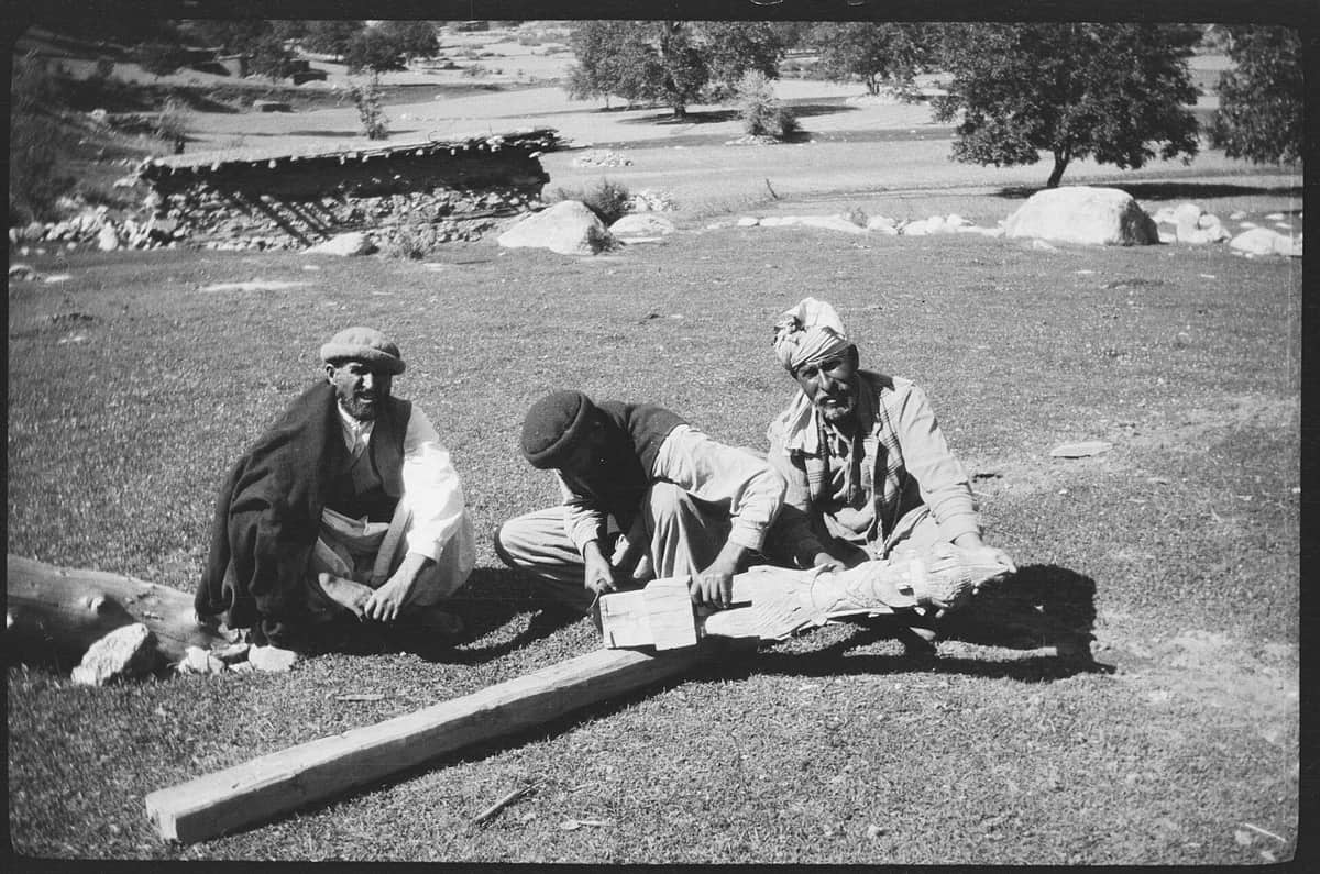 A 1929 photograph of three men sitting on grass around a long timber, one of them carving the head of a wooden ancestor figure