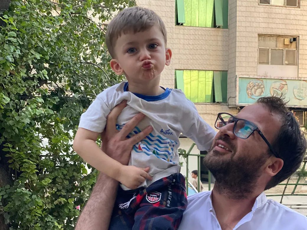 یوسف امیری، نمایندهٔ نورستانی در افغانستان، با عینک و پیراهن سفید، پسر خردسالش عبدالله امیری را در کوچه زیر درختی بالا گرفته است