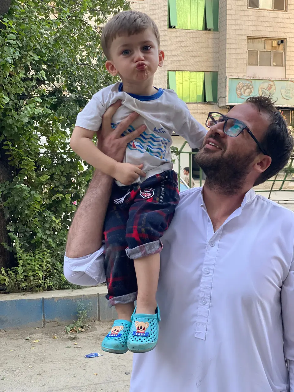 Yousuf Amiri, in glasses and a white shirt, smiling up at his young son Abdullah Amiri, who sits on his arm in a white T-shirt and blue sandals and puckers his lips for the camera, in a street with a tree and apartment blocks behind