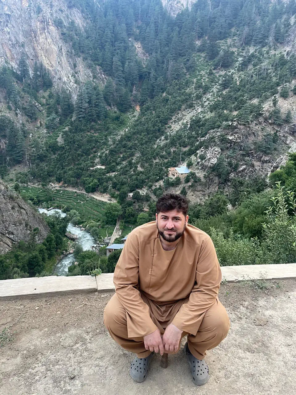 Zabiullah Amiri, director of Nuristani, crouching on a wall above the Sewketra valley in Waygal district, a turquoise river, terraced fields and a timber house far below and cedar forest on the mountainside
