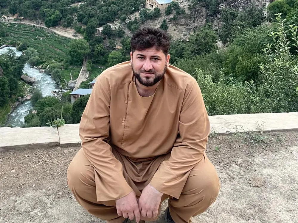 ذبیحالله امیری، مدیر نورستانی، بر دیواری بالای درهٔ سیوکترهٔ ولسوالی وایگل، با دریای فیروزهای، کشتزارهای پلهای و خانهٔ چوبی در پایین و جنگل ارچه بر دامنه