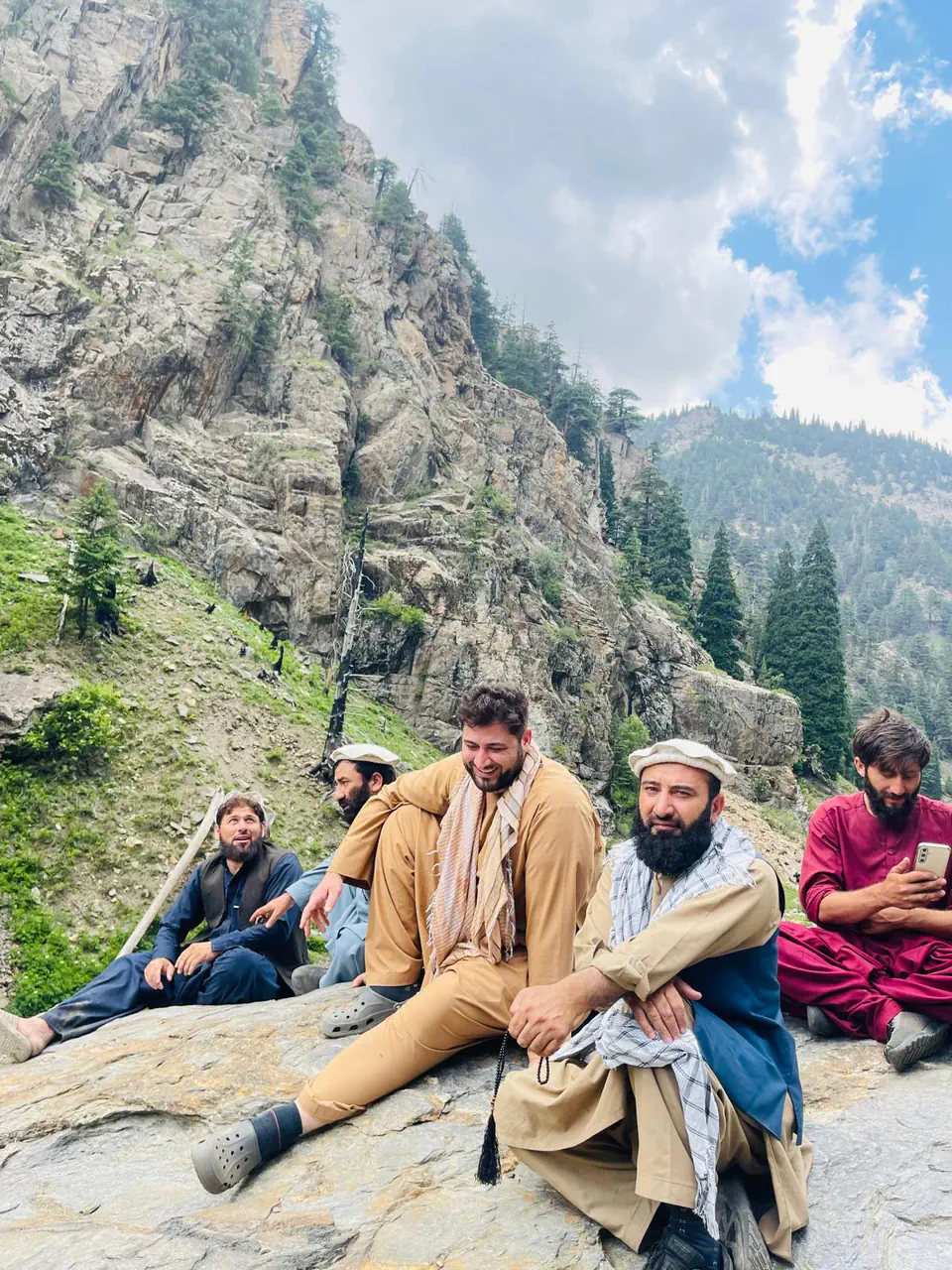 Zabiullah Amiri laughing with friends and family seated on a rock slab in Waygal, cliffs and pine forest rising behind, one man holding prayer beads